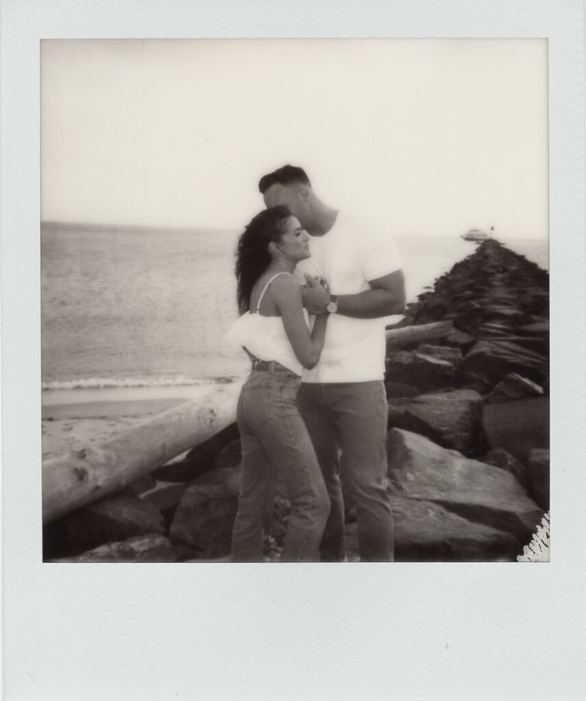Black-and-white Polaroid of the couple embracing beside driftwood and the stone jetty at their Cape May beach engagement session
